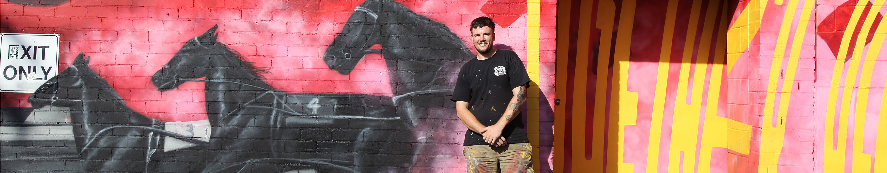 A smiling man with clothes covered in paint stands in front of a bright artwork featuring three black racing horses painted on an industrial building.