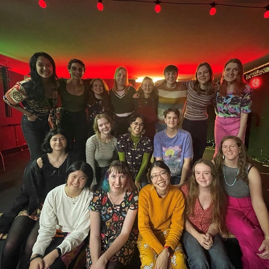 Group photo of Spectra Jazz, 18 young musicians standing and sitting in 3 rows in a red and green lit studio space, with a red light coming from the back of the room and red spot lights on a green hue at the front of the ceiling. The group are smiling towards the camera and all dressed casually. 