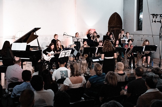 Spectra Jazz Orchestra playing on stage in white church-like space with white walls. The audience is visible in the foreground and the conductor is stood in front of the orechstra, dressed in black and with thier back to the audience. There is a pianist on the left with the remainder of the orchestra to the middle and right, all dressed in black.