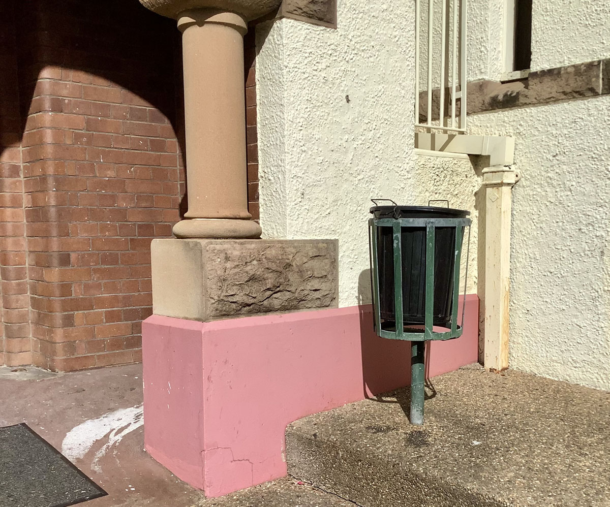 A fairly close view of an inner-suburban building showing a variety of textures: bricks, a sandstone column rising from a pink-painted smooth rendered wall, a white-painted wall with a rougher render, and a pebble-paved ground with a steel rubbish bin positioned above it.