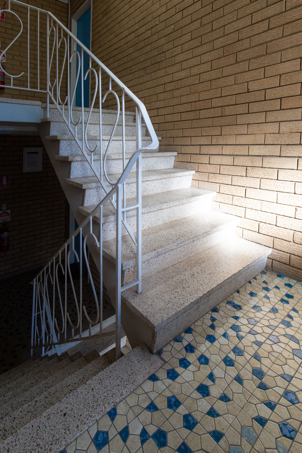 The stairwell of a walk-up apartment building. The lower and upper sections of the stairway meet at a blue-and-white-tiled landing, lit by evocative daylight from a window.