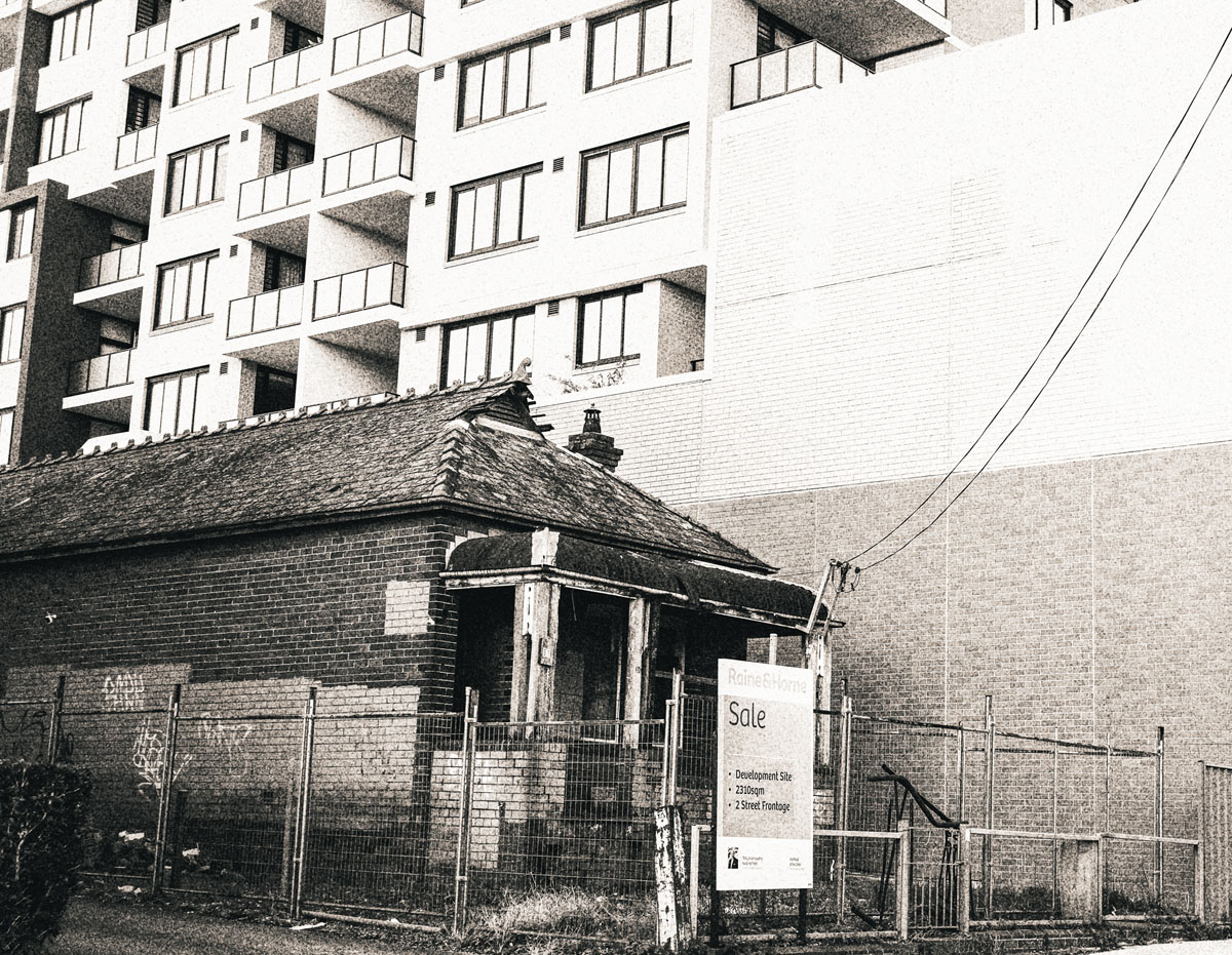 A dilapidated single-family cottage house, gated off and with a real estate SALE sign outside, stands directly alongside recently-built apartments next door.