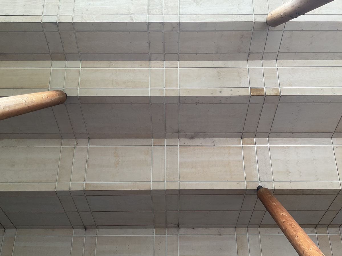 An abstract upward view of the concrete ceiling of a building with multiple ridges, with supports rising from the sides of the frame to meet it.