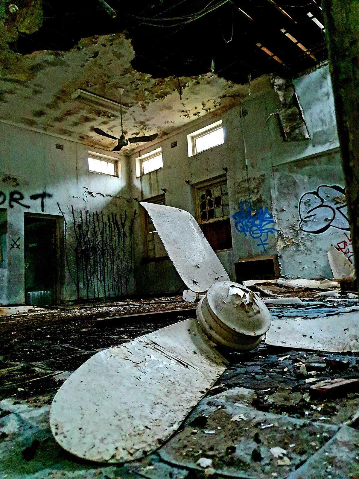 A room in a heavily dilapidated and abandoned building. A large unattached propeller sits among small detritus on the floor.