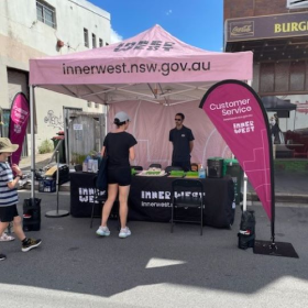 People standing in front of an information stall covered by a pink plastic marquee that sees 'Inner West'. A clear sky is behind them.