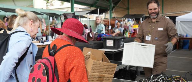 People browsing a market table one person wears a red ladybird shaped backpack. A man in an electricians uniform behind a trestle table is carrying a large white box
