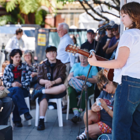 People sitting and standing on a street watching a performance. A musician in the foreground is playing a guitar