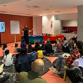 A group of adults and children sitting on a library floor listening attentively to a presenter standing at the front of the room talking in from of a screen
