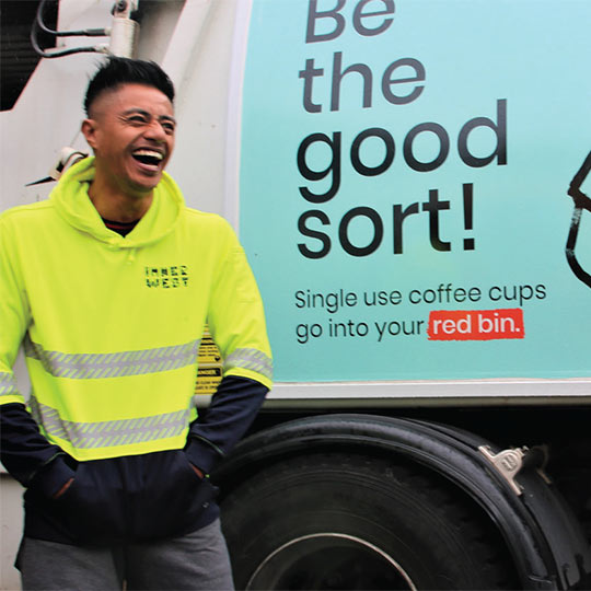 A resource recovery officer candidly laughing next to a garbage collection truck with 'Be the good sort! Single use coffee cups go into your red bin' emblazoned on the side.