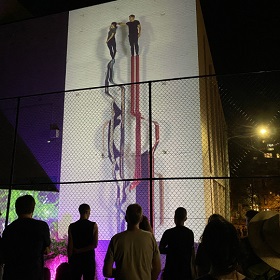 Photo of projection on a wall at night there are two ribbon like shapes running horizontally up the wall and two people standing at the top of each ribbon, a crowd looks up at the projections