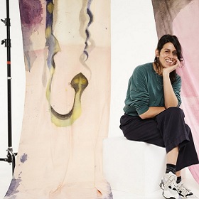 Photo of artist in a studio, in background is the artists large abstract painting of broad brush strokes, browns and pinks, artists sits perched cross legged in front of her artwork on a stool 