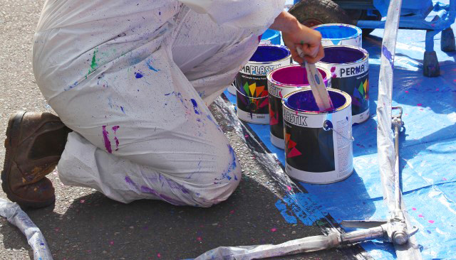 Photo of a painter in white overalls, only visible are the painters legs and they are surrounded by pots of paint - their overalls are covered in flecks of different paint