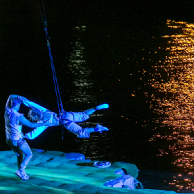 Nighttime photo of Legs on the Wall performers one on feet in harness the other lifted off ground, water reflections photo by Brett Boardman