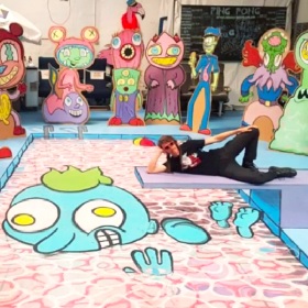 Photo of an artist lying on one side of the floor the floor is covered in an artwork of a cartoon character, the character has blue skin, green hair and a silly expression on its face, in the background are a life-size series of  similar style cartoon characters