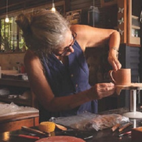 Person stands at a studio table and works with orange coloured clay their hands look like they are shaping a mug or jug. the person wears blue overalls and hair pulled up and glasses. There are tools on the table