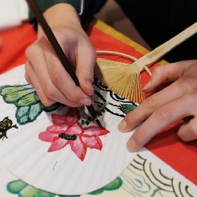 Photo of hands painting a fan with watercolours, the fan looks like a trasitional paper fan and the drawing is of a pink flower