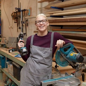 Photo of an artist in the Join The Dots studio work room she hold a drill and is surrounded by woodworking materials, she wears a marrone jumper and a green work apron