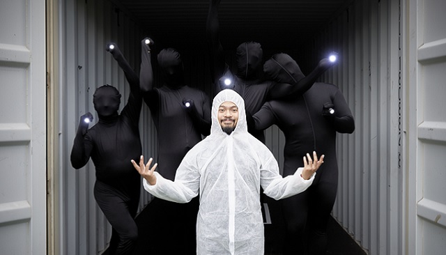 Photo of Shaun Parker and Co performance, person at forefront of photo wears a hazmat hooded white suit, they smile and have a black goatee, in the background performers with black body suits covering their head and face hold up little hand held torches and the effect is like looking at glowing balls being held in the air by the person at the front of the photo, in the background you can see part of a grey shipping container that the performers are standing in 