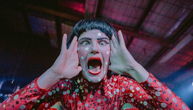 Photo of woman with white sequin dots on her face, short black fringe, woman has hands to the sides of her face in a outraged or shocked expression, her mouth makes a large O shape , she is wearing a red shiny long sleeve top also with white and silver sequins