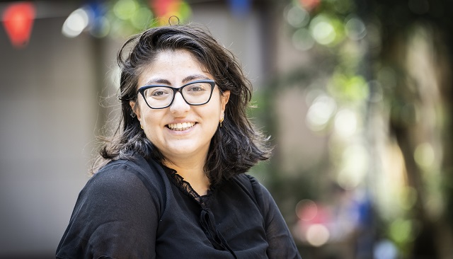 Photo of Eda smilking at the camera, they have thik shoulder lenght brown hair, dark rimmed glasses and a black blouse