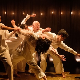 Photo of dancers mid performance they bend and lean over each other wearing white clothing, the lighthing is slightly warm and sepia and on the wall are little lights that seem to be attached t0 the wall in a dash shape