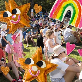 Phpoto of a a large crowd of lots of children and adults sitting and standing on the grass, cut out style illustration has been placed ontop of the photo and includes a rainbow, two gold glittery stars with smiley faces abd two smaller gold glittery hearts