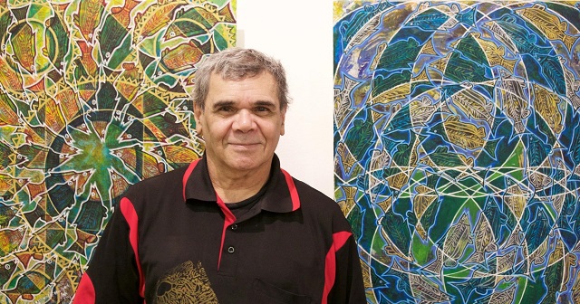 Portrait of Jeffrey Samuels by Sharon Hickey, Jeffrey stands in front of two large paintings on the left painting there is a star shape in the centre and white outlines show many fish shapes all heading to the centre star, the more you look the more you see thebackground colours are green, yellow, orange, brown and dark green-blue, the painting on the right is similar but its colours are blue, green and yellow - the fish are ore in a repetitive circular shape and there are white overlapping circles. Jeffrey stands in front of these paintings smiling with his mouth shut, he has a very friendlyface and grey short hair, he wear a black polo short sleeve shirt with red highlights and a gold emblem