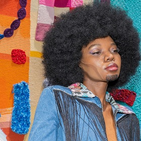 Photo of a person with a blue 70s style upcycled denim jacket with black long fringe, they have a beutiful large round afro and eyemakeup, there is a chiseld cheek contouring makeup on the persons cheek and they have a black short goatee style beard. Behind the person in a crochet panneld artwork