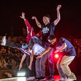 Photo of four performers on a street stage they are in various acrobatic / popping poses, arm up, legs out, bodies hunched, they are wearing street style clothing and its nighttime, you can see a crowd in the background and there are red lights shining onto the performers