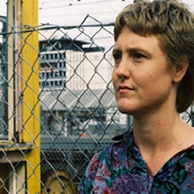 Colour Photograph of woman with short blond hair in front of a wire fence, train station in background