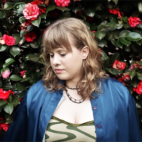 Photo of person standing infront of a flowering bush, it has large pink blooms that look like camerilas. The person has a rich bluey silver jacket, a nose ring, shoulder length sandy blonde hair and eye makep, they look away from the camera