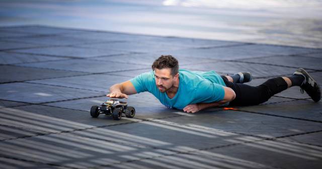 A person lies on soft form tiles they wear a aqua blue tshirt and black pants and have a dark short beard and dark hair, their hand hovers over a remote control car