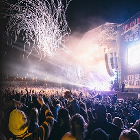 Photo of evening live performance show, white light fireworks are seen in top left of image and a stage in background, there is a large audience in the foreground and we see their backs mostly as they face the stage