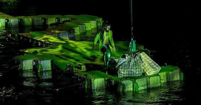 Legs on the Wall performer stand on a plontoon ontop of water with jacket, long hair, and bare chest wearing a suit jacket and shiny leggings, person has beard and circular chest tattoo. There are two large laundry bags attached to a pulley