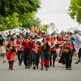 Marching band on the road wearing red, many brass intruments can be seen and there is a crowd of people following