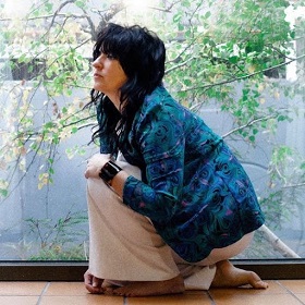 Photo of woman crouching on knee, dark hair long hair with fringe, black arm cuff, blue jacket, white flares, barefoot