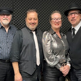 Four performers pose, smiling for the camera all wear black, white, grey or silver