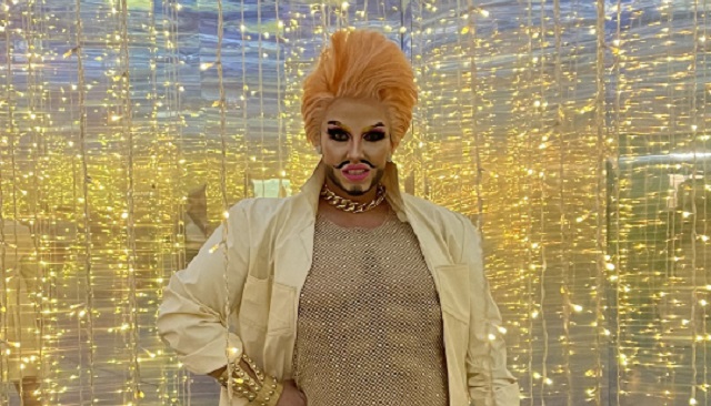 A Drag King dressed fabulously in golden glitter stands in a shiny room