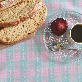 Table with check print tablecloth, round loaf of bread, slices, plum half, black coffee, crystal plate