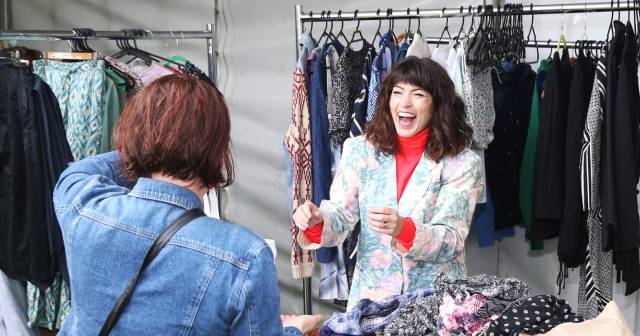 Woman standing among clothing speaking with woman who\