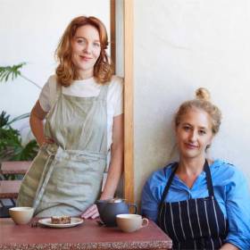 One woman standing one woman sitting both wearing aprons