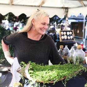 Smiling woman leans over green plants at a stall