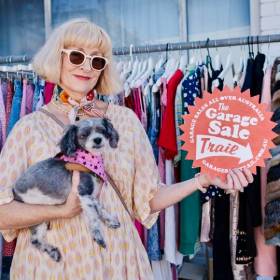 Woman with grey hair holding a small dog stands in front of a rack of clothes and holds a sign saying Garage Sale Trail