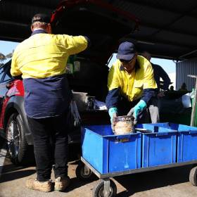 Two mean unloading old paint tins from the boot of a car into a blue container