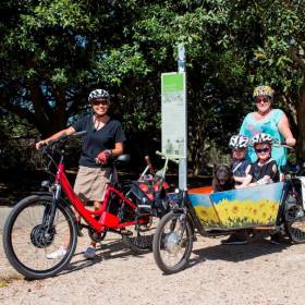 One adult with two children in a cargo bike and another adult with a bike standing behind them