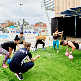 Group of people in a circle outdoors on astro turf doing various workouts under supervision by a trainer