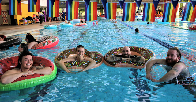 Four people in inflatable rings in a swimming pool with rainbow flag bunting hanging above the pool