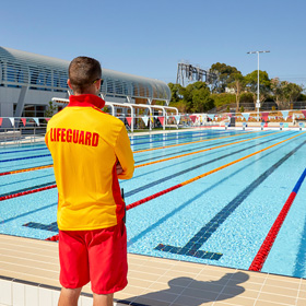 Man wearing yellow and red shirt that reads "Lifeguard" looking over a pool