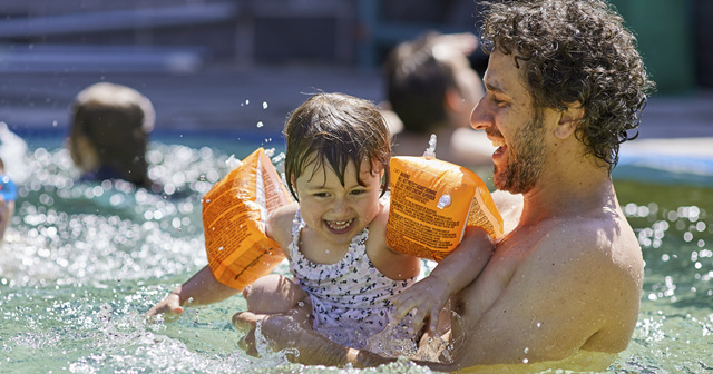 Man in mushroom pool with little girl wearing orange water wings splashing and smiling