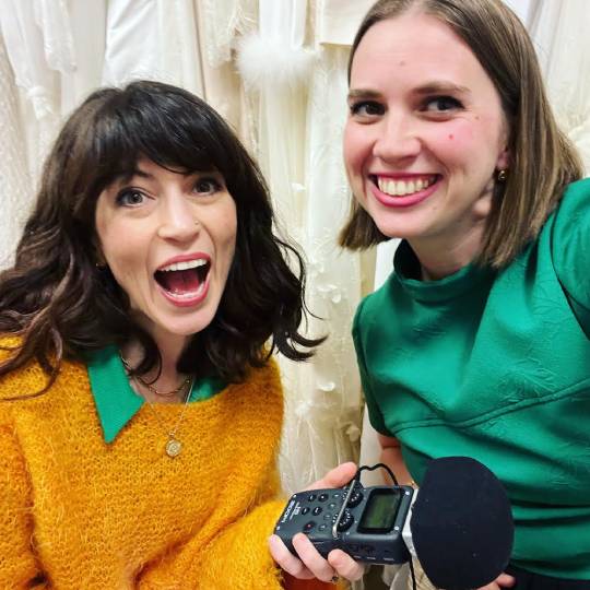 Two smiling people huddled close in front of a wall of fabric. One is holding a microphone.